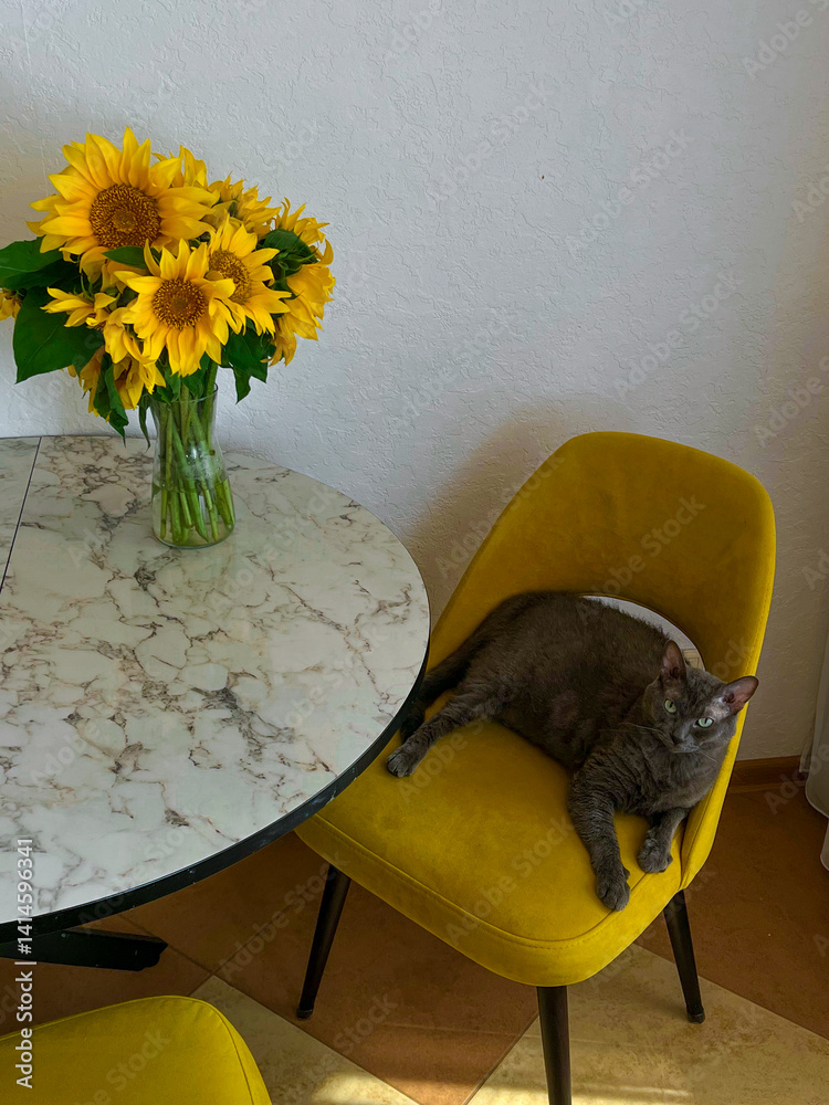 A grey cat is lying on a yellow armchair near a table with a bouquet of sunflowers