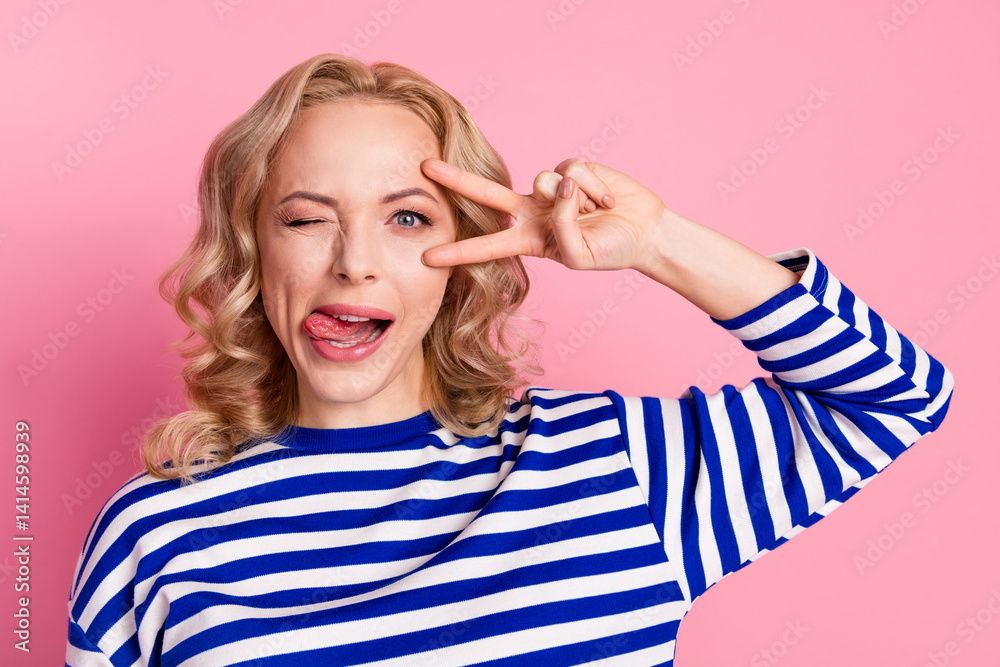 Fototapeta premium Young woman in striped pullover making a playful peace sign gesture with her hand in front of a pink background