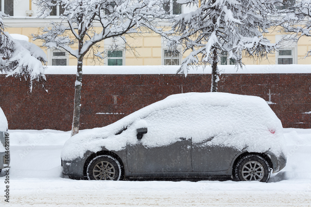 A snow-covered car on a snowy street. Heavy snowfall in the city. There is a lot of snow on the hood, windows and roof of a parked car. Snowdrifts on the roadsides. Cold winter weather.