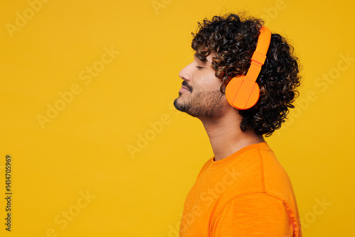 Side profile view young smiling happy Indian man he wears orange t-shirt casual clothes listen to music in headphones close up isolated on plain yellow background studio portrait. Lifestyle concept.