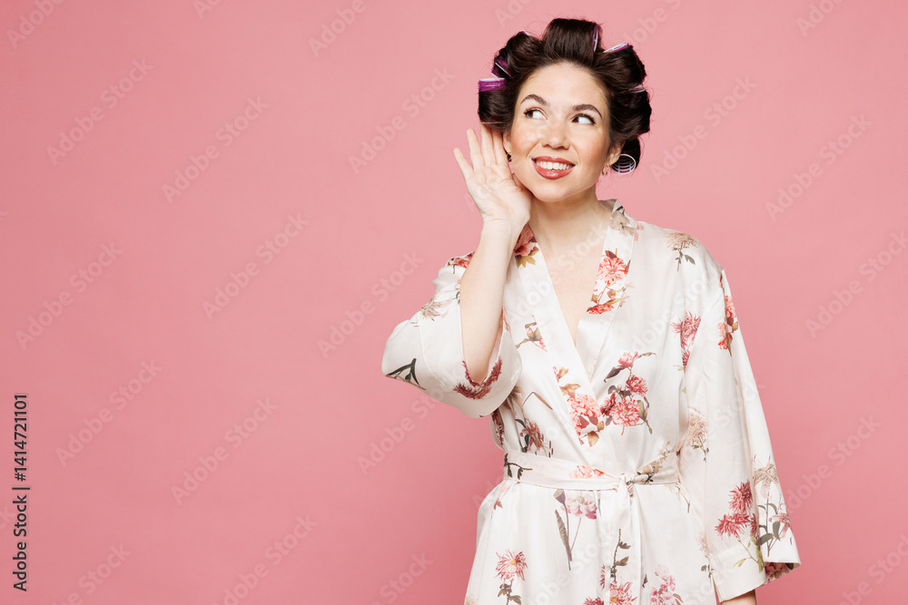 Young housewife housekeeper woman she wear white robe hair rollers try to hear you overhear listening intently isolated on pastel light pink color background studio portrait. Morning routine concept. Young housewife housekeeper woman she wear white robe hair rollers try to hear you overhear listening intently isolated on pastel light pink color background studio portrait. Morning routine concept.