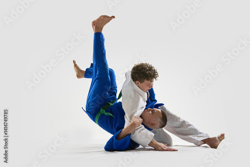 Young judoka in white holds opponent in blue during intense ground control, demonstrating control, and strength against white background. Concept of victory, martial arts, combat sport, competition.