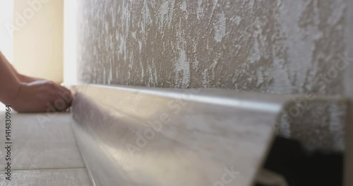Experienced builder carefully applying plastic skirting board to wall using masking tape during meticulous apartment renovation process, demonstrating professional craftsmanship and precision