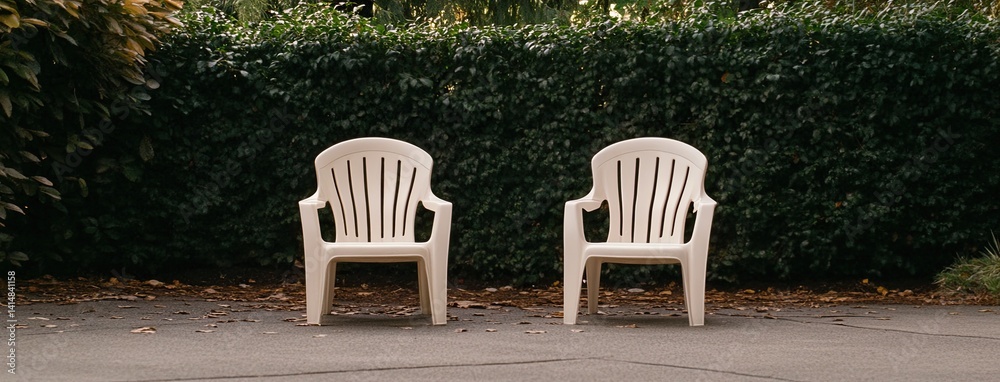 Two white plastic chairs sit peacefully in a garden, surrounded by lush green hedges and grass under warm summer light.