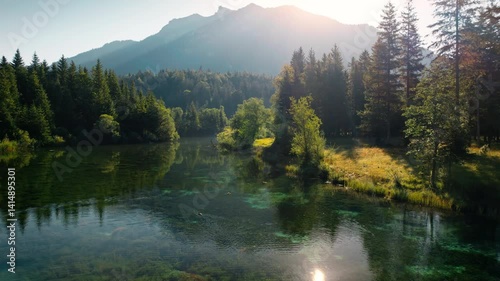 Flying over painterly lake surrounded with conifer forests.
With the sun shining over the mountains, shot in Bavaria, Germany.
