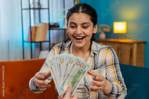 Indian young woman experiencing sudden happiness, joyfully celebrating on sofa receiving plenty of cash dollars. Arabian girl sincerely delighted, smiling widely, enthusiastically showing victory win