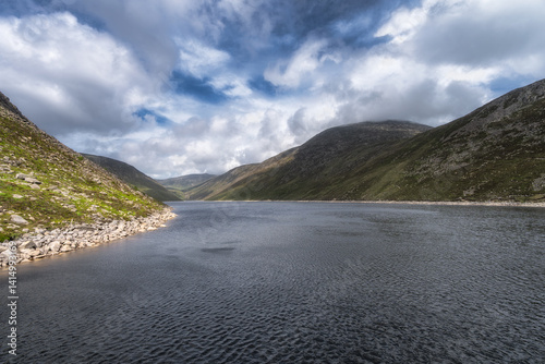 A serene mountain lake, Ben Crom Reservoir, beautifully situated among rolling green hills of Mourn Mountains, captured beneath a dramatic cloudy sky, ideal for nature enthusiasts, Northern Ireland