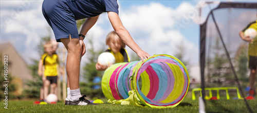 Fototapeta Naklejka Na Ścianę i Meble -  A coach sets up a colorful tunnel during an outdoor kids' sports activity. Children in yellow jerseys participate eagerly, holding a ball and preparing for a fun obstacle course