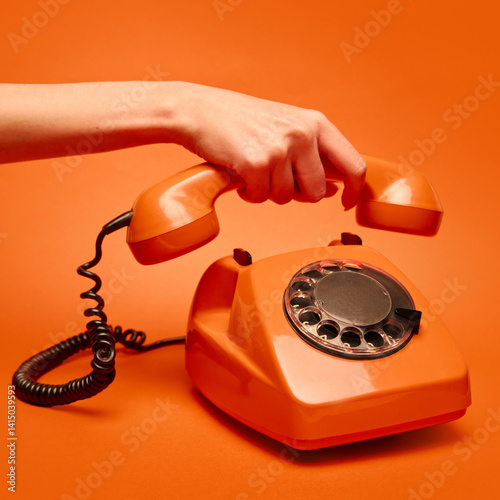 Obraz na płótnie Hand lifts the receiver of a vintage orange rotary dial telephone against a matching orange backdrop. Concept of retro, vintage, technology, pop art. Suitable for creative visuals, posters w salonie