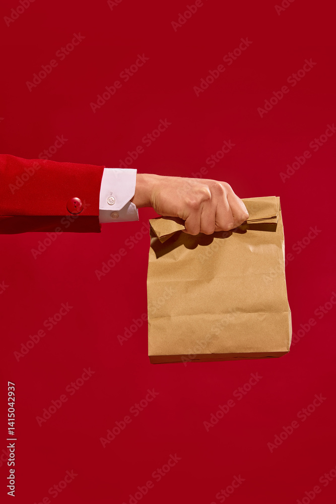 Hand holding kraft paper bag against bold red background. Express delivery with eco-friendly packaging. Concept of courier, branding, takeaway. Food delivery, sustainable brand ads, express service