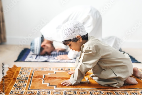 A Muslim father and his young son, both dressed in traditional attire, engage in prayer on prayer mats. The father prays while the son learns the importance of prayer. photo