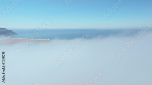 Aerial view of Rauðasandur, Iceland, where golden sands meet turquoise waters, framed by dramatic cliffs and low-hanging clouds under a radiant blue sky