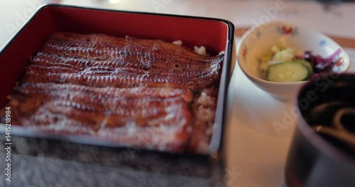 Close-up shot of traditional Japanese unagi donburi with glazed grilled eel over rice in a lacquered box as the camera makes a slow semicircular movement above the dish