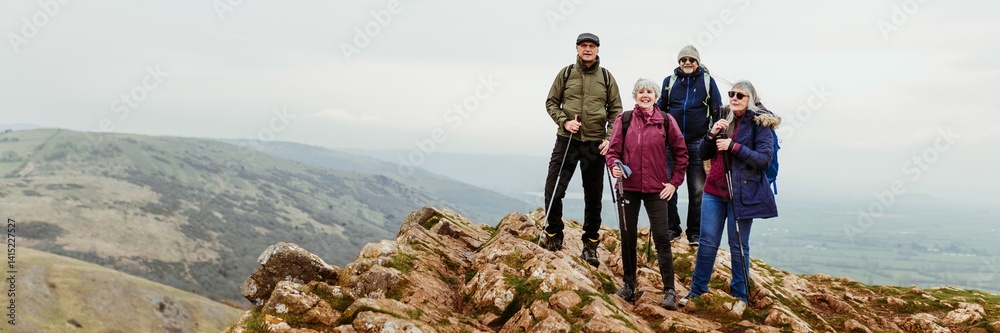 A diverse senior group of four hikers stand on a rocky peak. The hikers, dressed for the outdoors, enjoy view of the mountains and valleys. Happy senior group with hiking dressed at a rocky peak