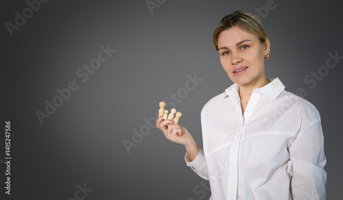 The image depicts a woman wearing a white shirt with buttons. She is holding a small group of wooden figures in her hand. She is looking away and smiling. The background is a solid gray with space for