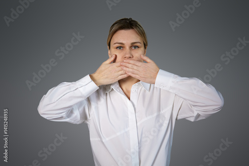 A woman on a gray background in a white shirt covered her mouth with her hands, silence, gray background. Blonde girl on gray background in white shirt keeping mouth closed, conceptual photography