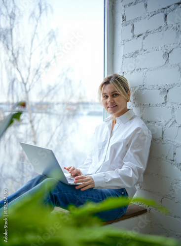 A young woman in a white button-down shirt and blue jeans sits by the window using a laptop. The girl is smiling, freelancing, business, teaching. Behind her back is a window and to her right is a