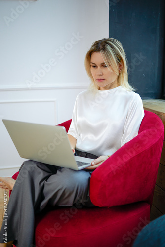 A beautiful woman sits in a bright red chair working on a laptop. She is wearing a white shirt and gray pants. In the background is a light colored wall with space for copy. A cubicle in an office or