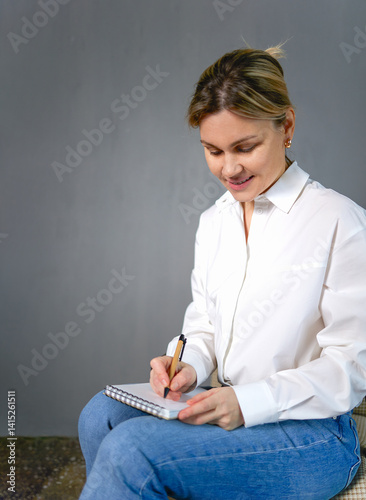 A woman in a white shirt and blue jeans sits writing in a small notebook with a pen. She is looking down at the notepad, a business, a teacher or a student. The background is a monochrome gray wall