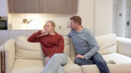 Apologetic regretful man pleads with upset distant girlfriend on couch. Emotional strain, trust shattered, relationship conflict, heartache, reconciliation attempt, love tested, emotional distance