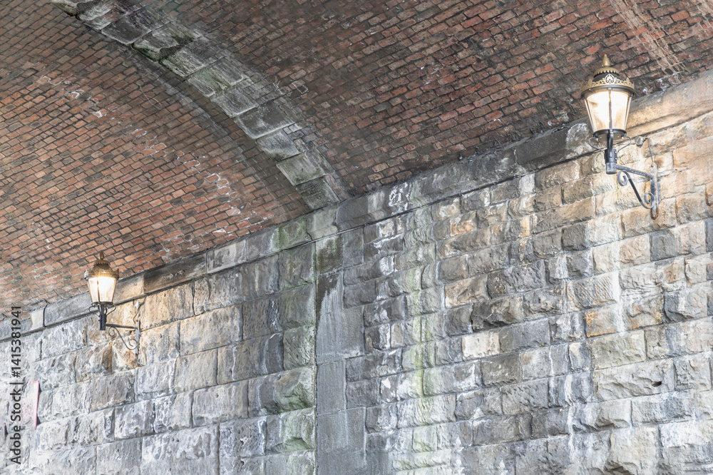 Two lantern lighting a spot on stone wall and red brick ceiling in tunnel under the high level bridge. An curved brick tunnel with lamps under the bridge, use it as your Wallpaper, Poster and Space.