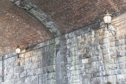 Obraz na płótnie Two lantern lighting a spot on stone wall and red brick ceiling in tunnel under the high level bridge. An curved brick tunnel with lamps under the bridge, use it as your Wallpaper, Poster and Space. w salonie