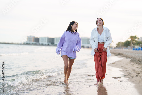 Two Female Friends Walking Happily on a Sandy Beach at Sunset in Relaxed Casual Outfits