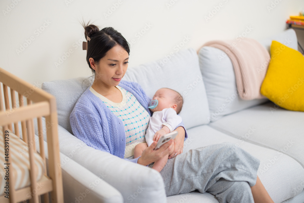 Mother using phone while holding sleeping baby on sofa