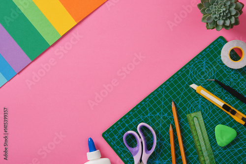 Supplies for paper art hobby: scissors, craft knife, pencil, eraser, cutting mat, colored paper on pink background. Copyspace. Paper crafts work table flatlay.