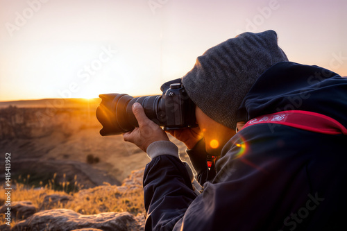The image shows a person wearing a beanie and a jacket, holding a camera with a lens, taking a photo outdoors during sunset