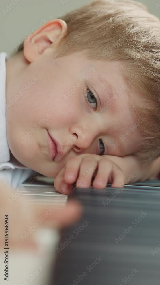 Cute blond boy rests head on piano keyboard. Sad child moves fingers along keys playing lazily. Sleepy kid learns to play musical instrument closeup