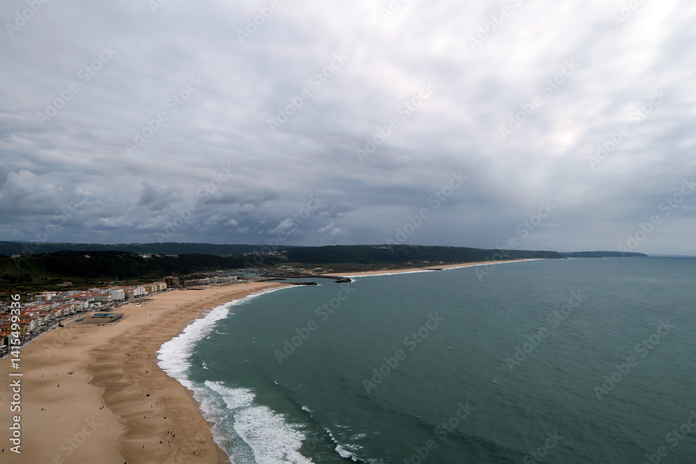 Fototapeta premium Panoramic view of a curved beach flanked by a coastal town and backed by green hills, under a cloudy sky stretching out over the ocean, in Nazaré, Portugal. 