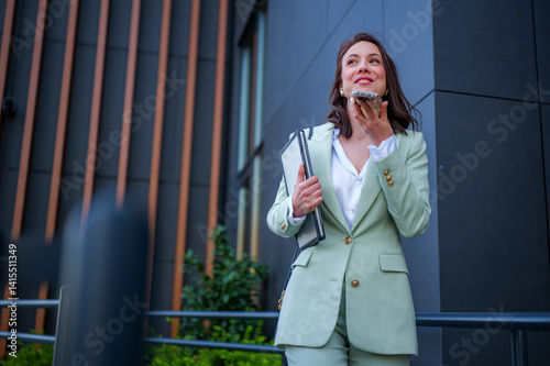 Smiling businesswoman wearing suit holding folder and recording voice message on smartphone while walking outside office building after work