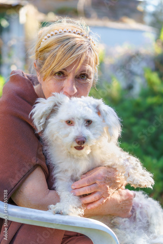 An elderly blonde-haired woman sits on a chair in the garden with her Bichon Bolognese dog, hugging and kissing it while both look at the camera.