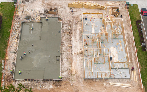 Workers smooth and polish concrete slab floor while other construct stud framework for new house