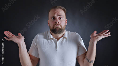 Фотография Man with a beard and tattoo, wearing a white shirt, raises his hands in confusion and disbelief