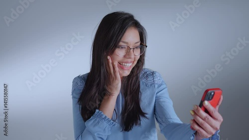 Woman gazing at her red phone, smiling, and touching her face, exuding joy and excitement.