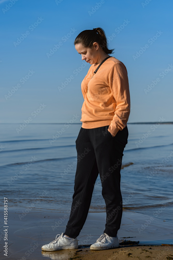 Pensive middle-aged woman in casual orange sweater and dark trousers enjoys the peaceful evening on a sandy beach by the sea. Pensive middle-aged woman in casual orange sweater and dark trousers enjoys the peaceful evening on a sandy beach by the sea.