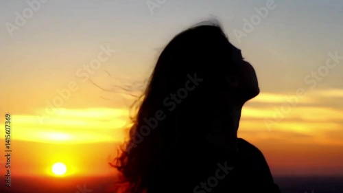 Silhouette of a woman gazing at vibrant sunset sky with wind blowing her hair, peaceful emotional moment captured at golden hour for meditation, freedom, nature and mood concepts.