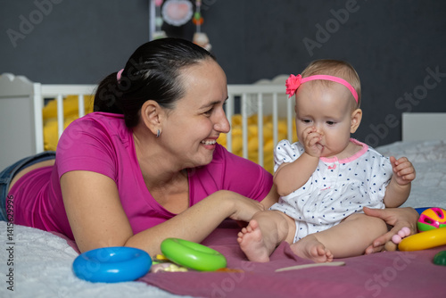 Mother and her adorable baby girl play together on a soft bed filled with colorful toys. Laughter fills the air as they share special bonding moments in their cozy home. 
