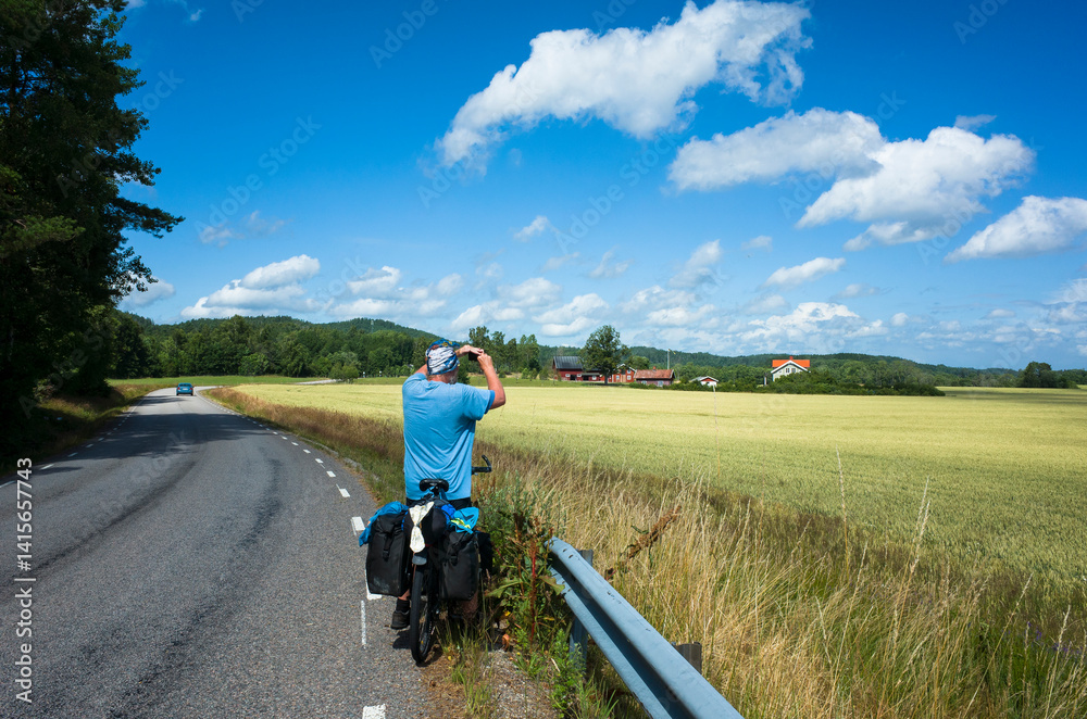 Fototapeta premium A bicycle traveler stops by the roadside in Västra Götaland County, Sweden, taking photo of the scenic countryside view. Golden fields and a bright summer sky create a peaceful rural landscape
