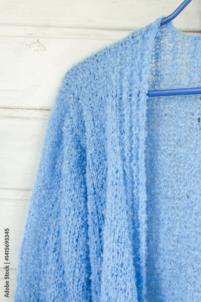 Blue cotton knitted cardigan on white wooden background. Soft blue knitted sweater on hanger in sunlight. Knitting pattern background. Summer knitted fashion. Cozy hobby. Craft product concept.