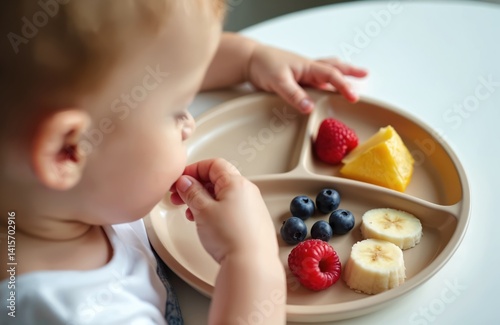 Toddler eats fruits from plate, close-up. Child hand taking food from beige divided plate. Kid aged one year, eating raspberries, blueberries, bananas. Baby enjoys healthy eating at home. Food for