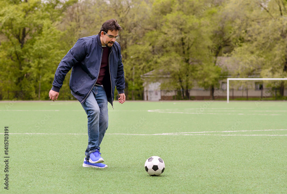 Obraz premium Middle-aged man in casual clothes playing soccer on green footbal field, ready to kick ball. Trees and goal posts in background. Active outdoor lifestyle and weekend recreation for adults. Copy space