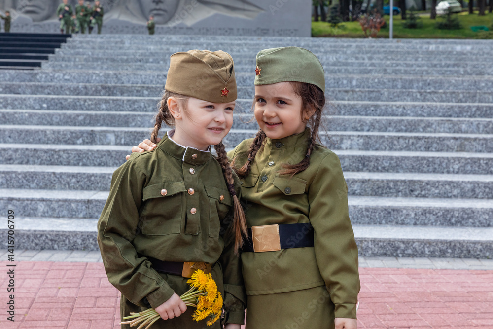 Patriotic education of children. Little girls with long braids in a military uniform, military cap, hold flowers in hands against the background of the memorial. Photo for the Victory Day on May 9
