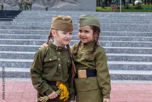 Obraz na płótnie Patriotic education of children. Little girls with long braids in a military uniform, military cap, hold flowers in hands against the background of the memorial. Photo for the Victory Day on May 9 w salonie