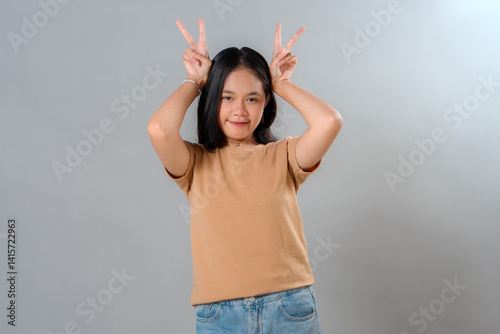 Happy young Asian woman smiling softly with a playful peace sign above her head, showing a cute and confident expression in a relaxed studio portrait against a plain background.