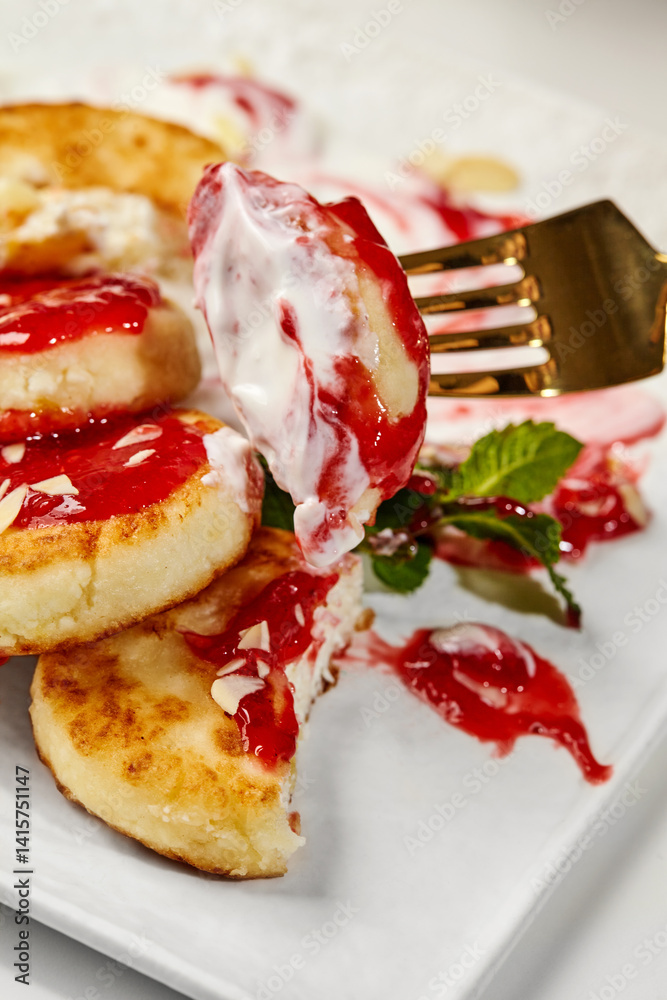Syrniki with strawberry jam, sour cream and almonds being eaten with golden fork
