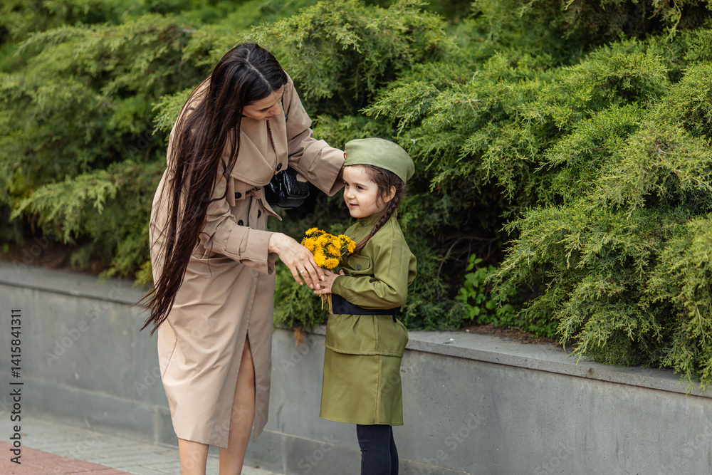 Patriotic education of children. Mother with little girl on Victory Day. Girl in a military uniform, military cap, holds flowers in hands near the memorial. Photo for the May 9 celebrations