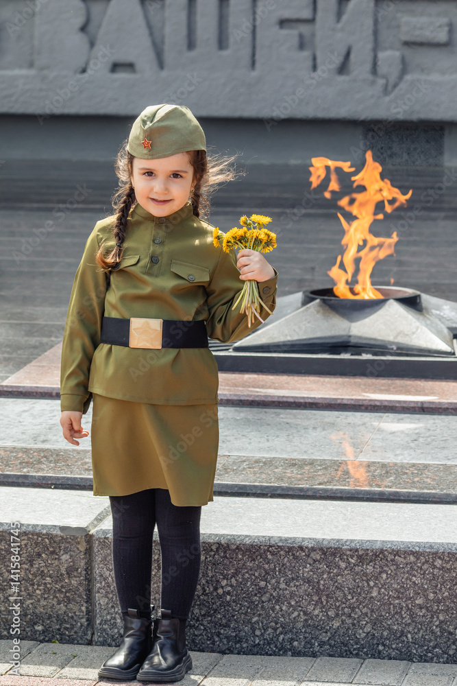 Patriotic education of children. Little girl with long braids in a military uniform, military cap, holds flowers in hands against the background of the memorial. Photo for the Victory Day on May 9
